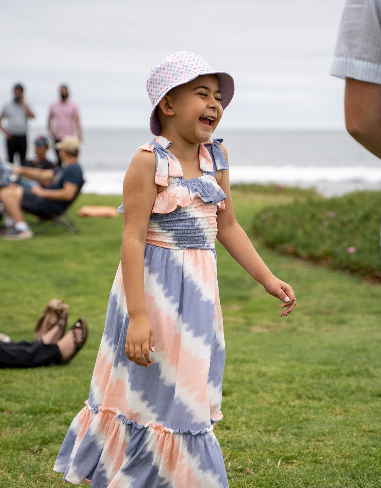 Girl with bucket hat laughs joyfully outdoors, ocean in the background. She wears a colorful dress.