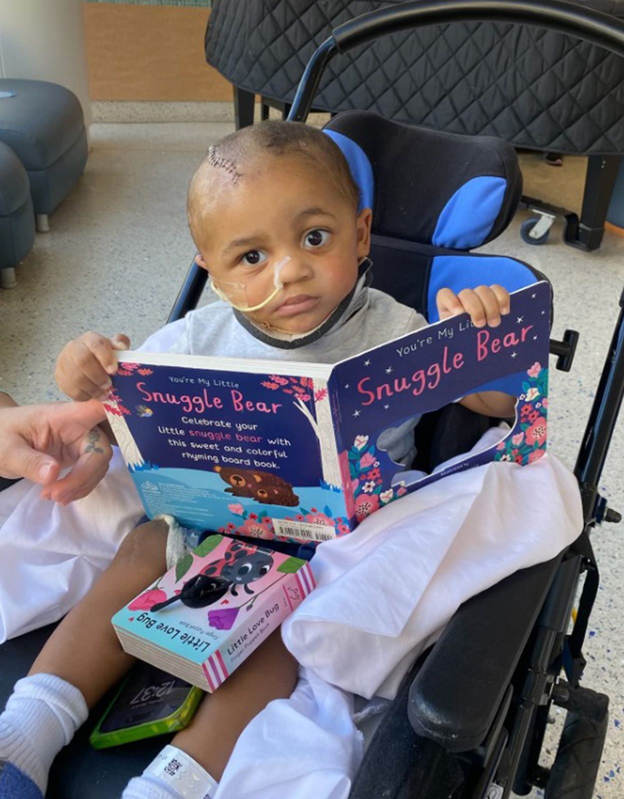 Young child in wheelchair reading a book. They have a nasal cannula, bald head, and thoughtful expression. Hospital setting.