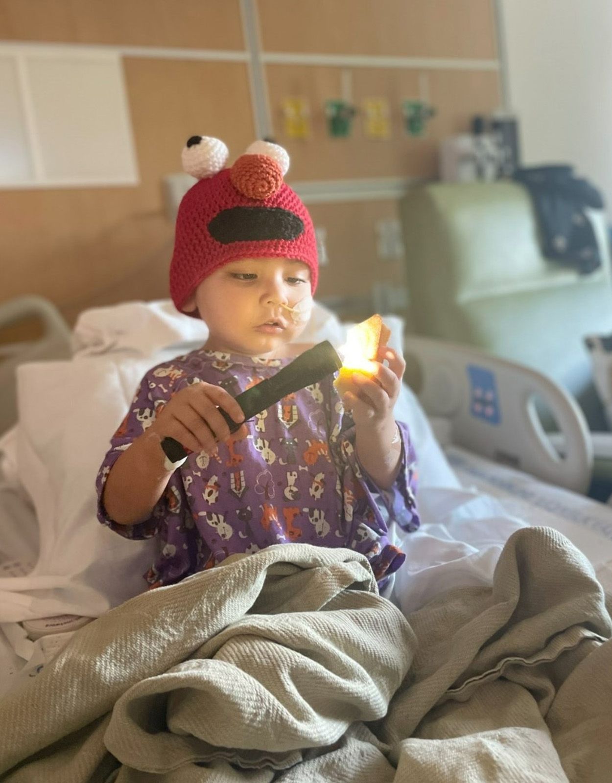 Child in Elmo hat in hospital bed, holding flashlight, focused expression.