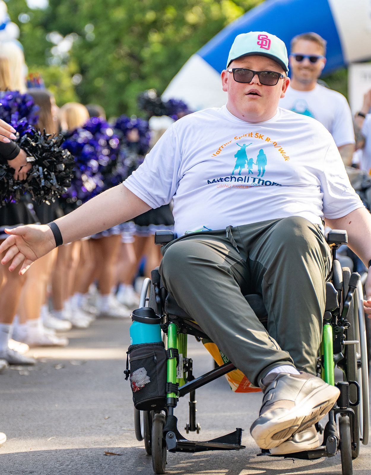 Man in wheelchair wearing a hat, white shirt, and sunglasses participating in a walk; cheerleaders in background.