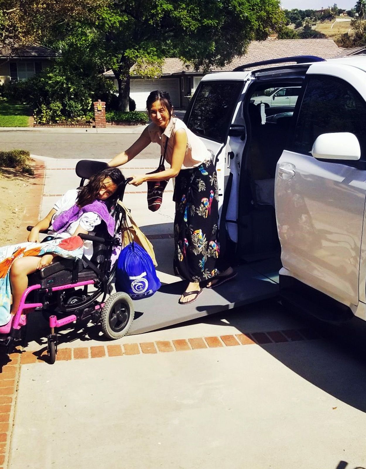 Woman helping a person in a wheelchair onto a white van with a ramp on a driveway.