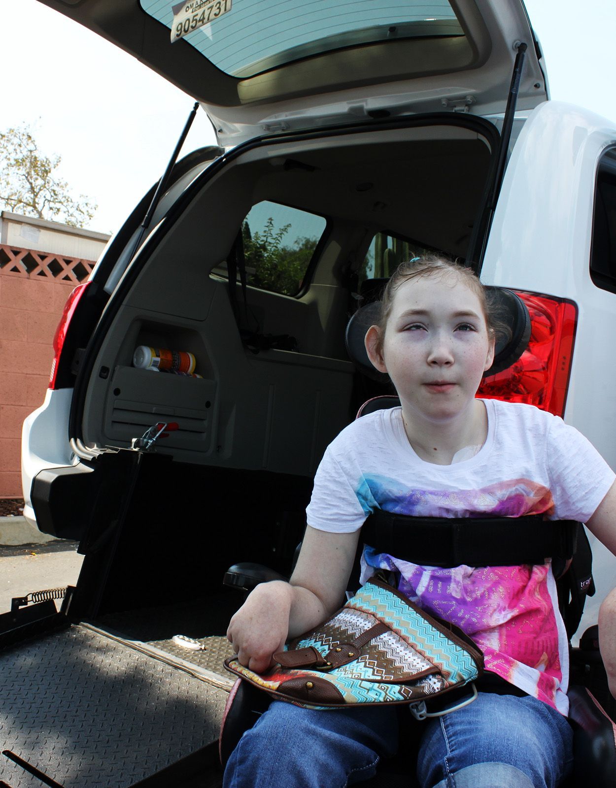 A girl in a wheelchair smiles near the open trunk of a white van with a ramp.