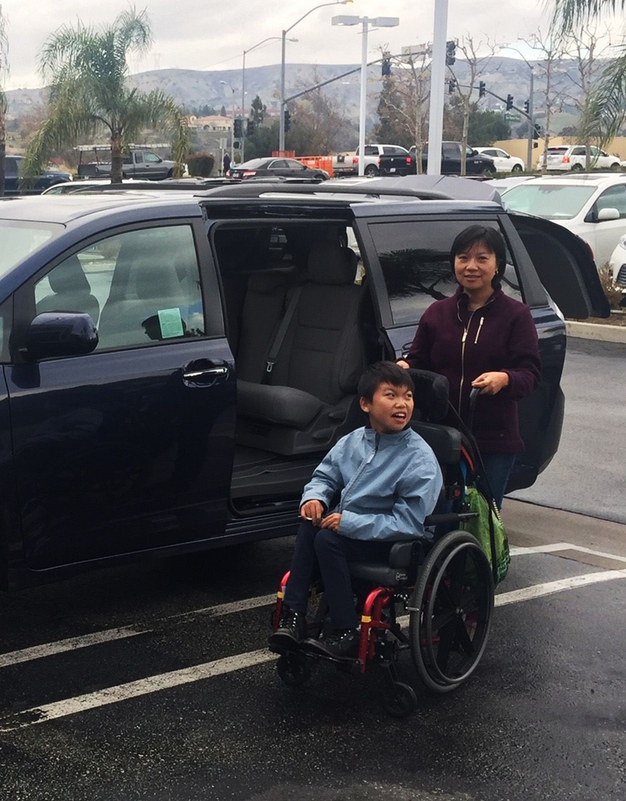 Woman and boy in a wheelchair next to a blue minivan with a ramp in a parking lot.