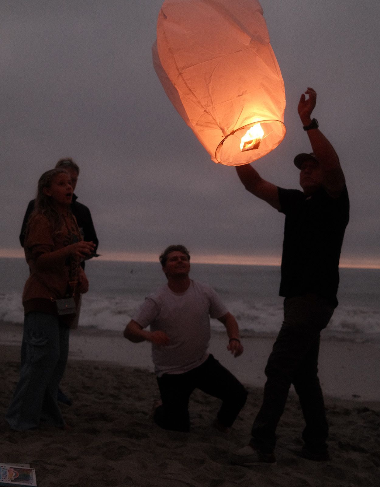 People launching a sky lantern on a beach at dusk. One man holds the lit lantern as another kneels; woman watches.