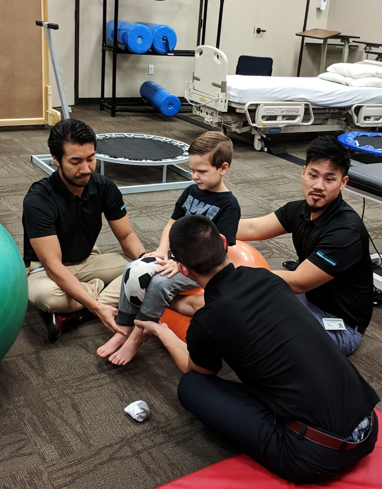 Four people helping a young boy sit on a ball; physical therapy room.