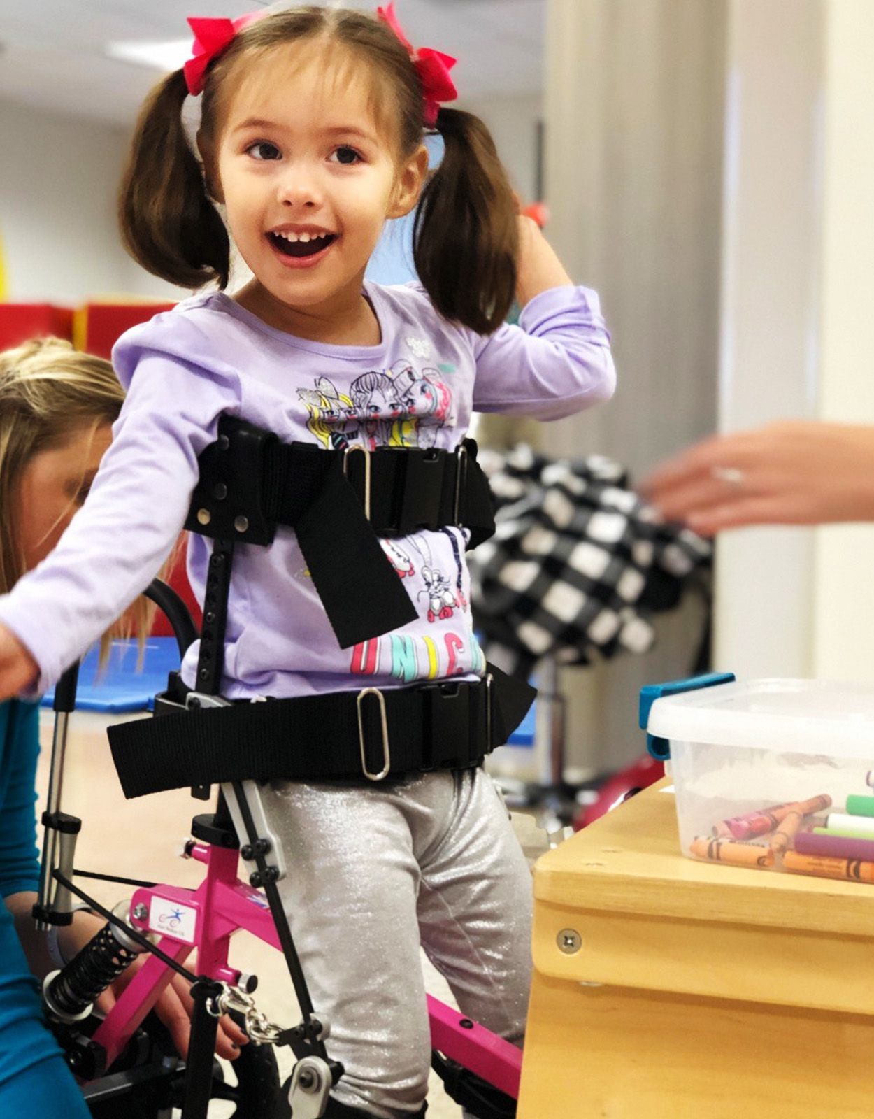 Smiling young girl with pigtails in a walker, wearing braces, excited in a playroom.