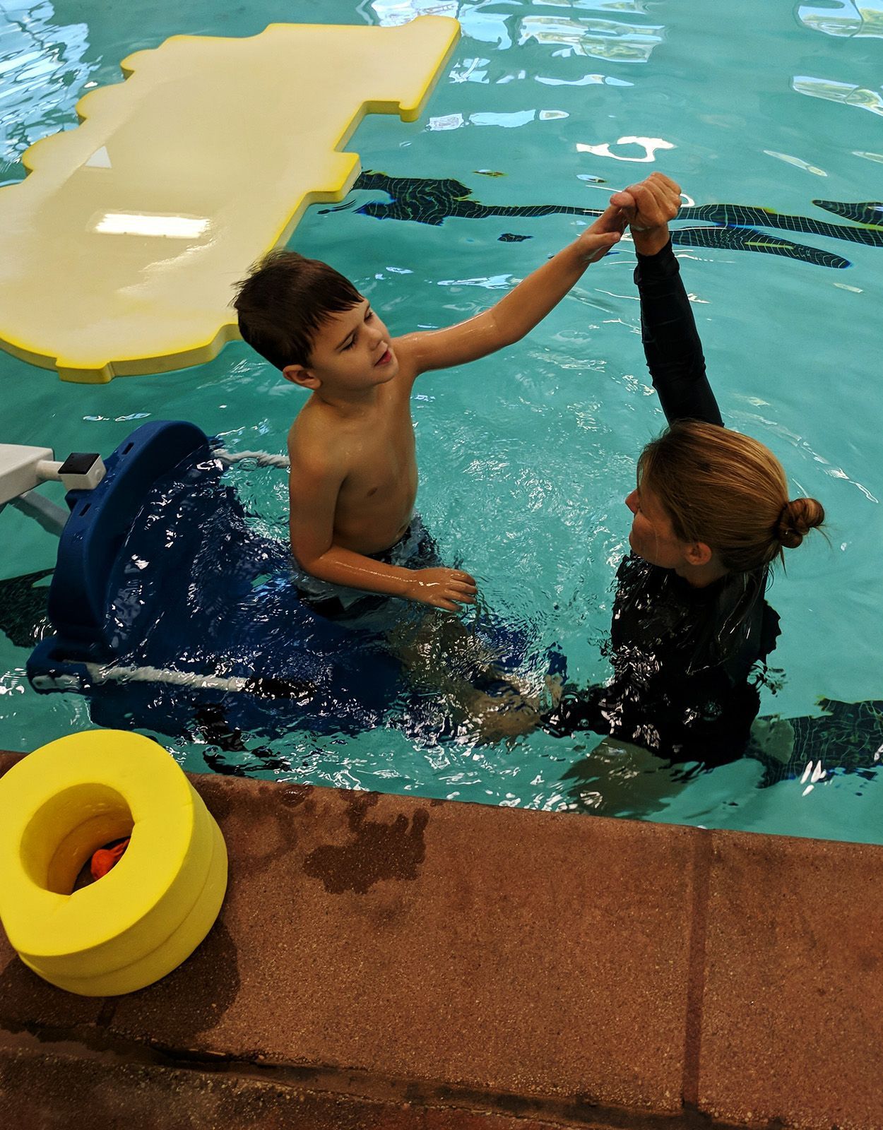 Boy in pool using a flotation device, reaching up to the instructor.