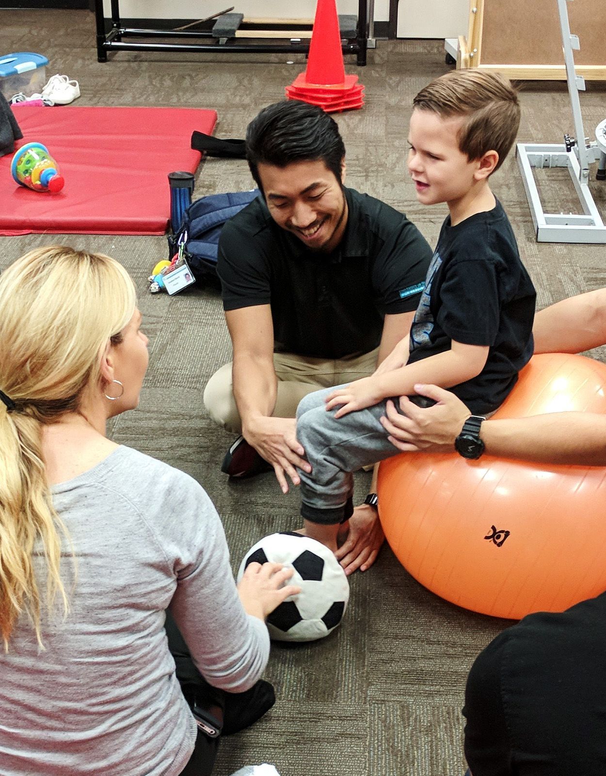 Therapist and child on exercise ball working with soccer ball. Smiling child, assisting therapist, and blonde-haired woman.