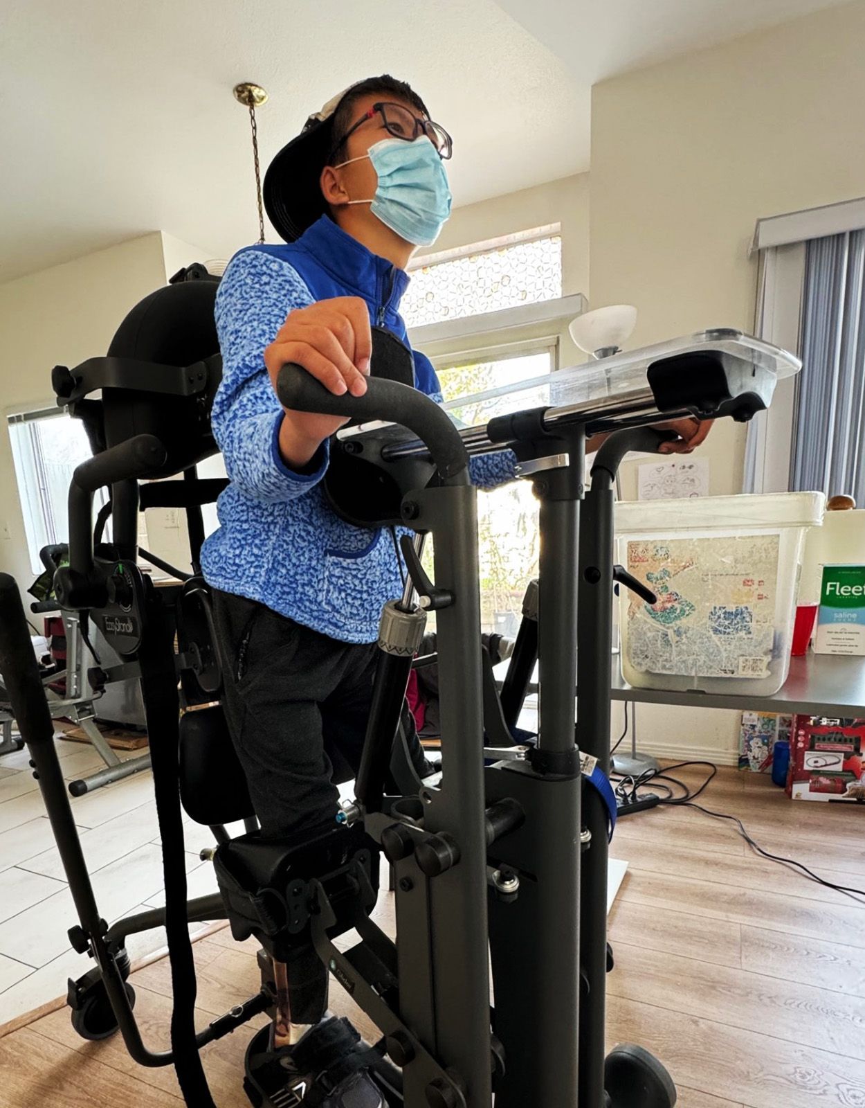 A person wearing a mask uses a standing frame indoors. They wear a blue jacket and look upwards.