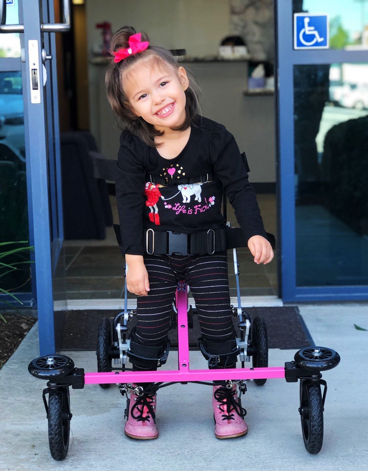 Girl smiling, using a pink walker outside a building. She is wearing a black shirt, striped pants, and pink shoes.