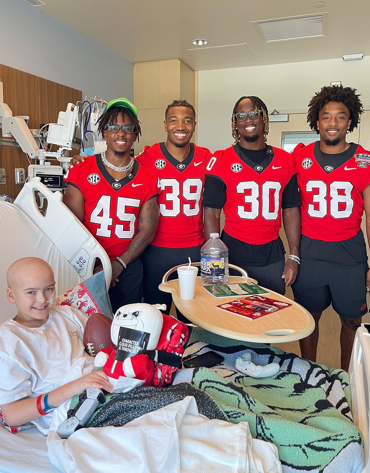 Four football players in red jerseys visit a smiling child in a hospital bed.
