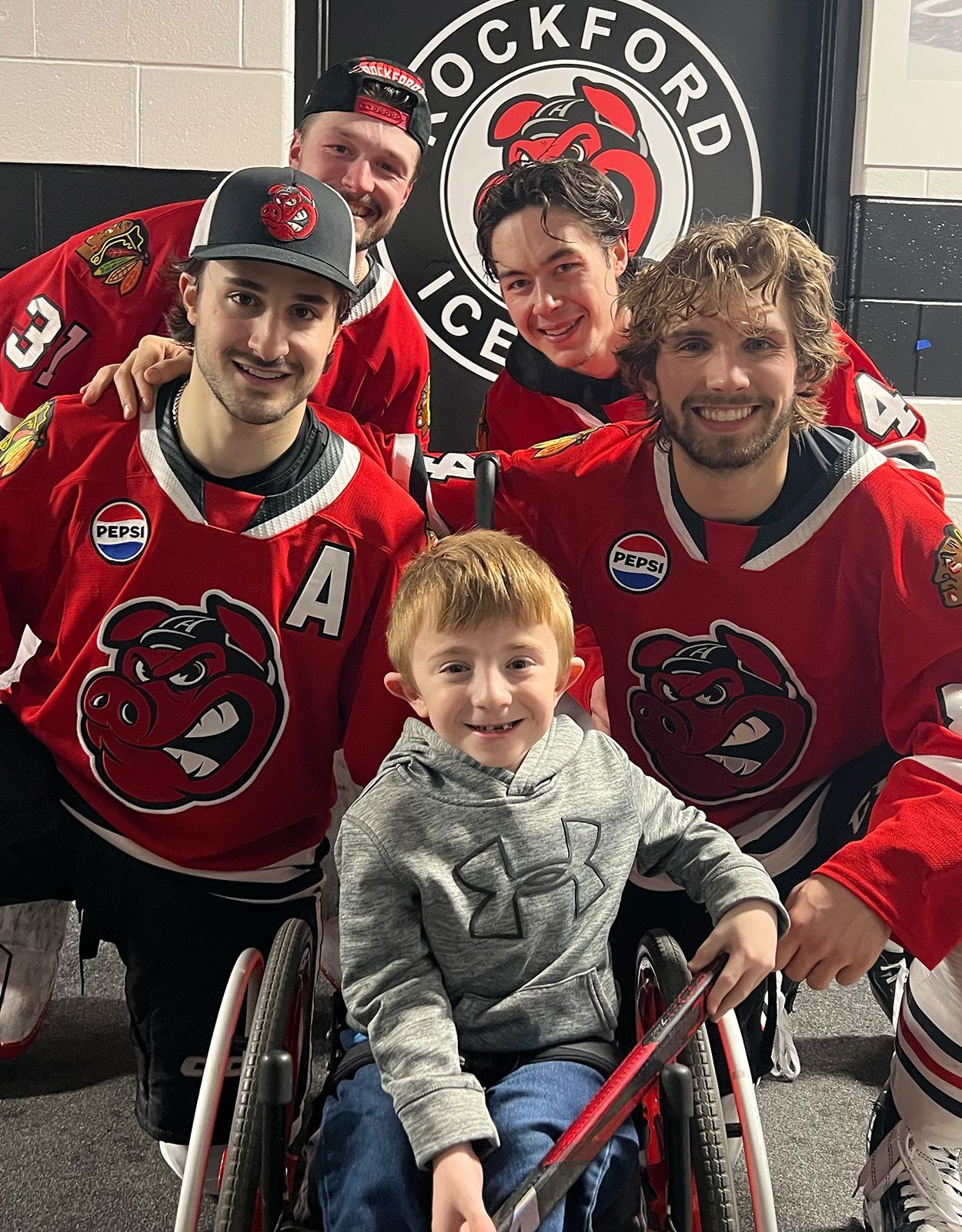 Hockey players in red jerseys pose with a smiling child in a wheelchair, in front of a Rockford IceHogs logo.
