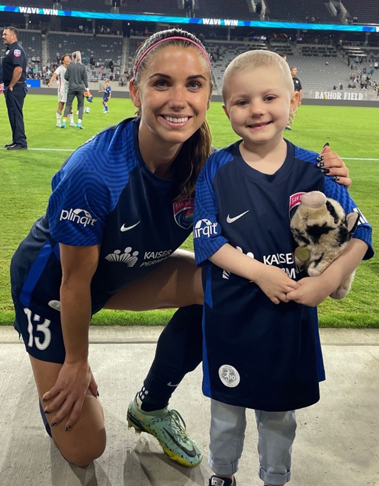 Soccer player Alex Morgan with a young child, both wearing blue jerseys, smiling at a soccer game.