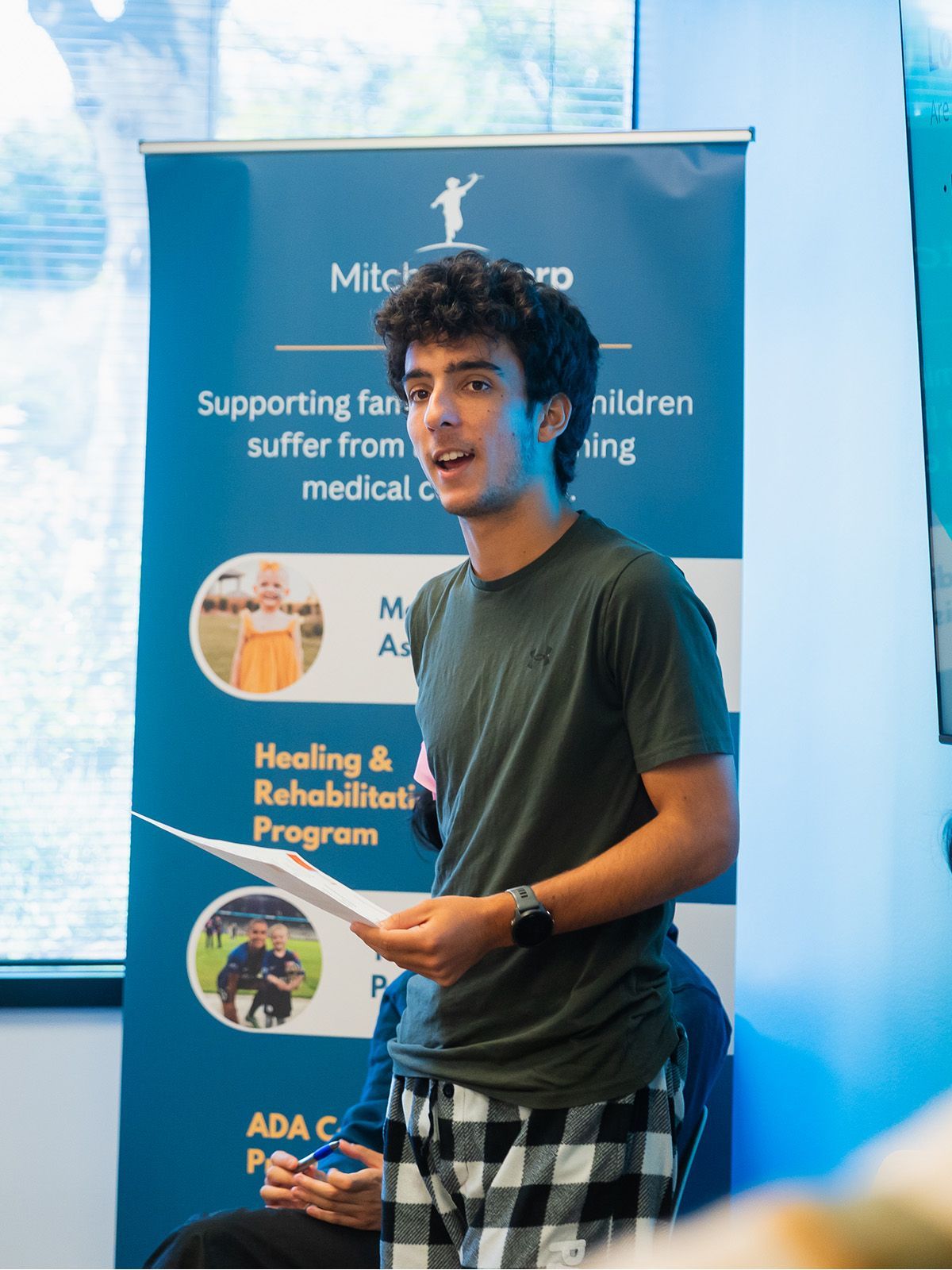 Young man speaking at event with banner in background.