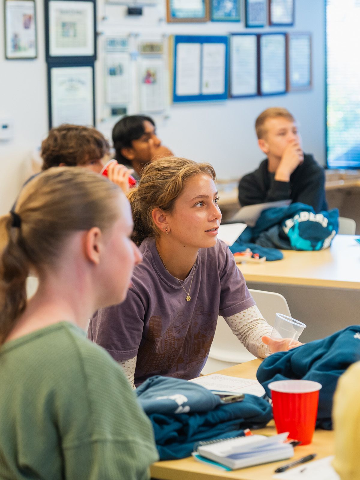 Students in a classroom, listening and engaged. Tables with papers and a red cup.
