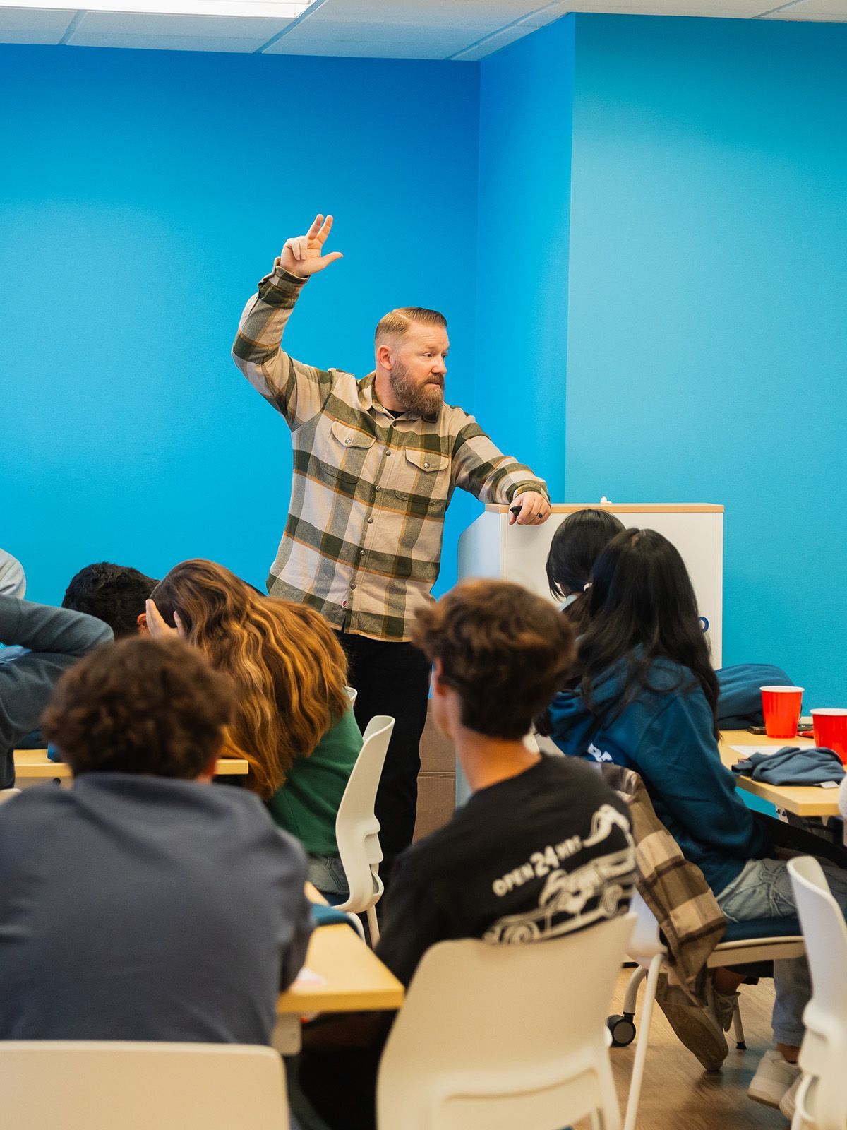 Man giving presentation to students in a blue room; he gestures with his hand.