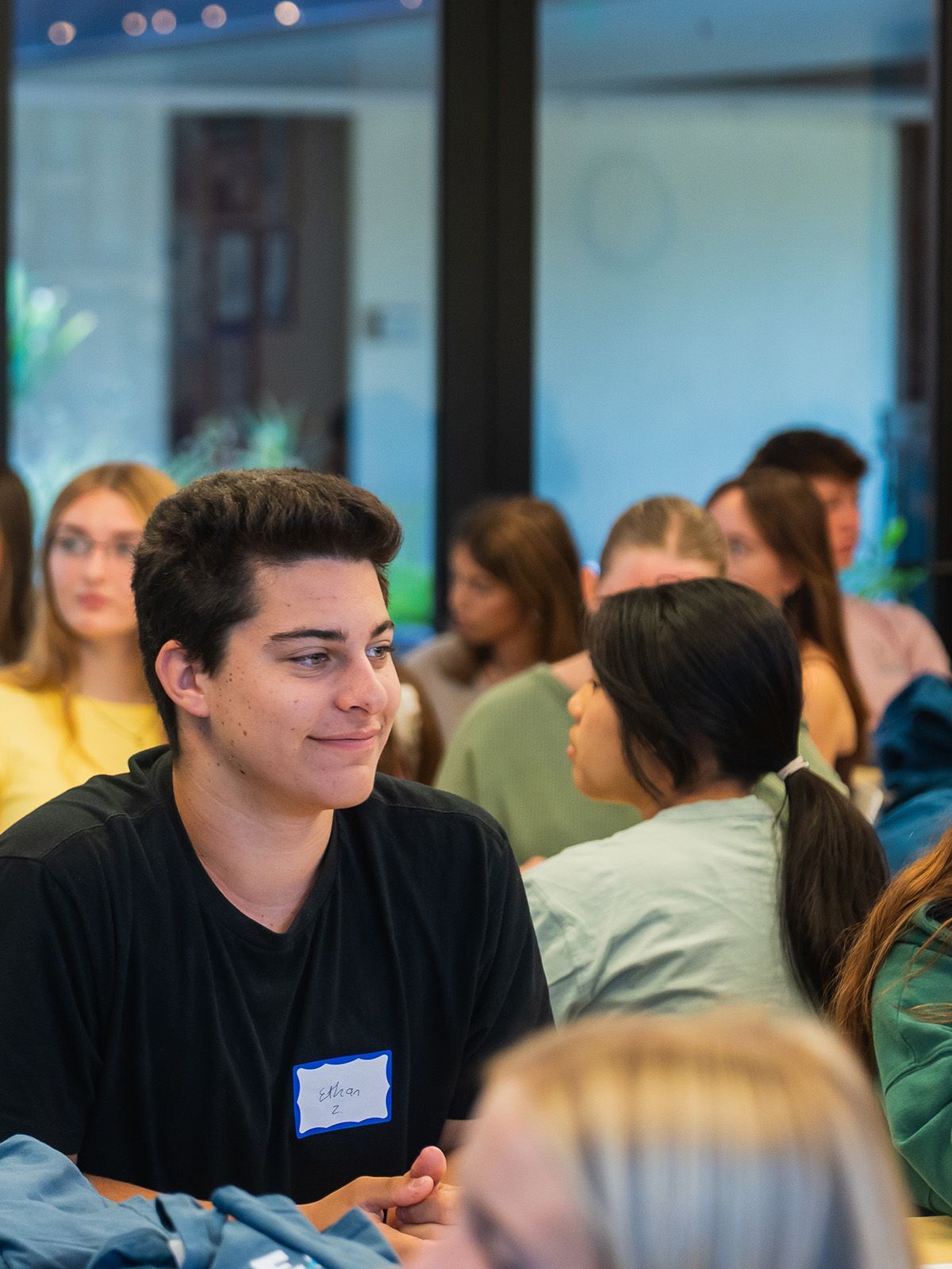 Young person in a black shirt smiles, seated in a room with other people.