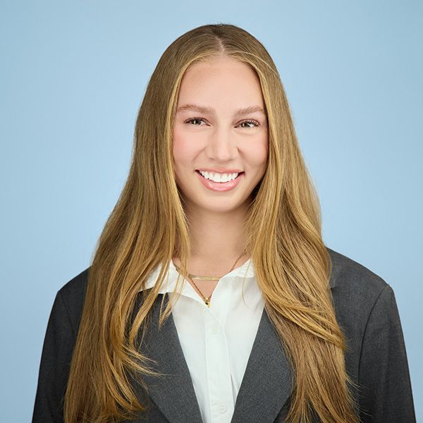 Woman with long wavy brown hair smiles at the camera, wearing a white shirt, against a green and red foliage backdrop.