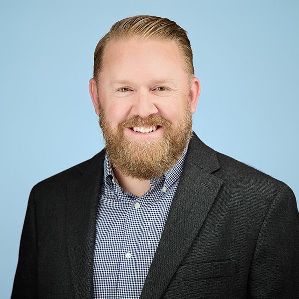 Man with red beard smiles, light skin, close up, in front of a basketball hoop.