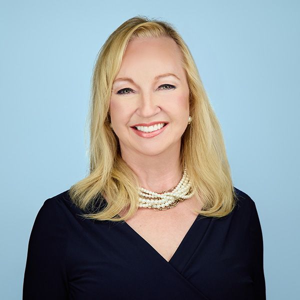 Blonde woman in a red blazer smiles, wearing a gold necklace, outdoors.