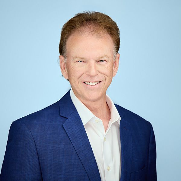Middle-aged man in a suit smiles at the camera; fair skin, light brown hair, neutral background.