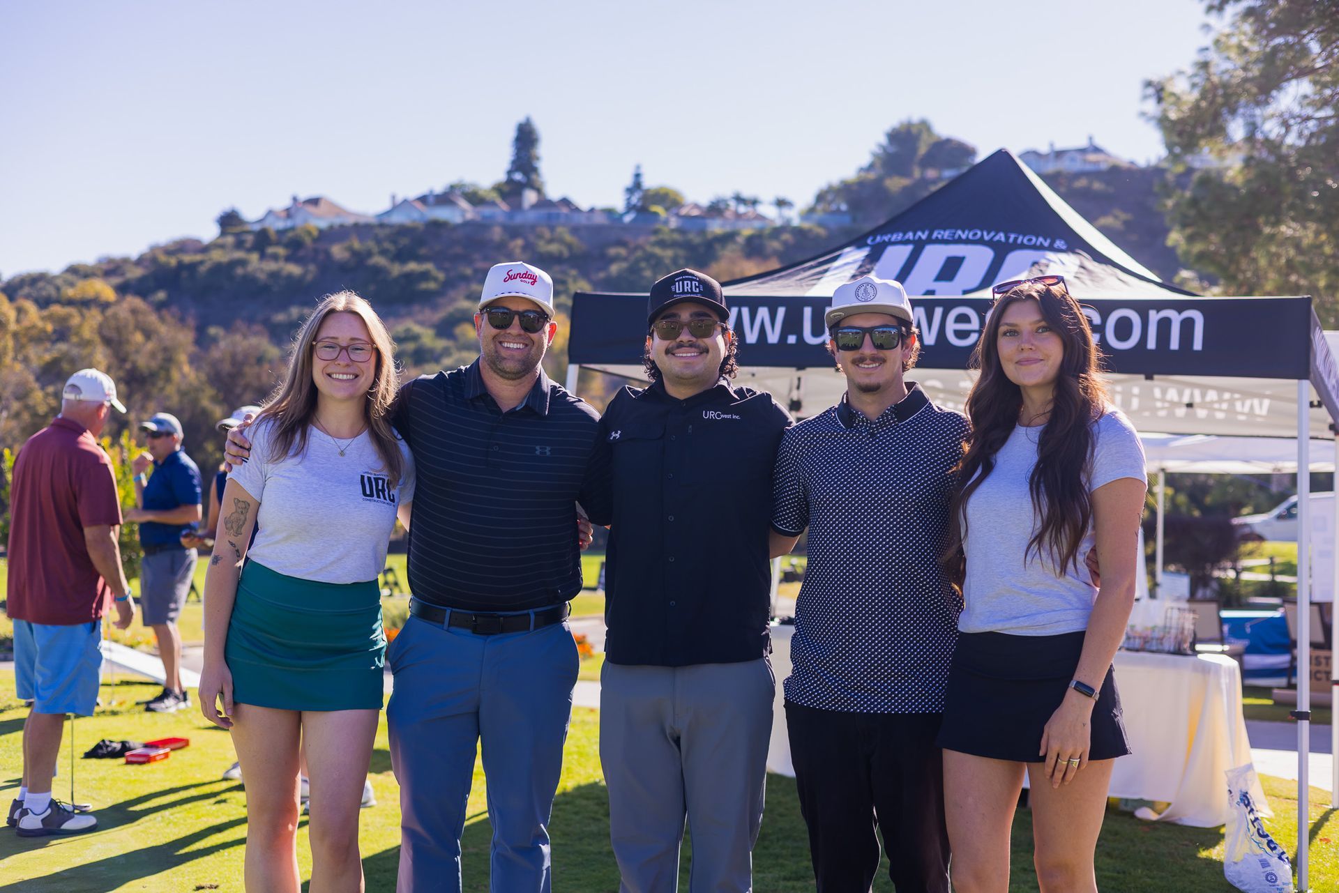 Five people posing for a photo on a golf course, with a tent and a hilly background.