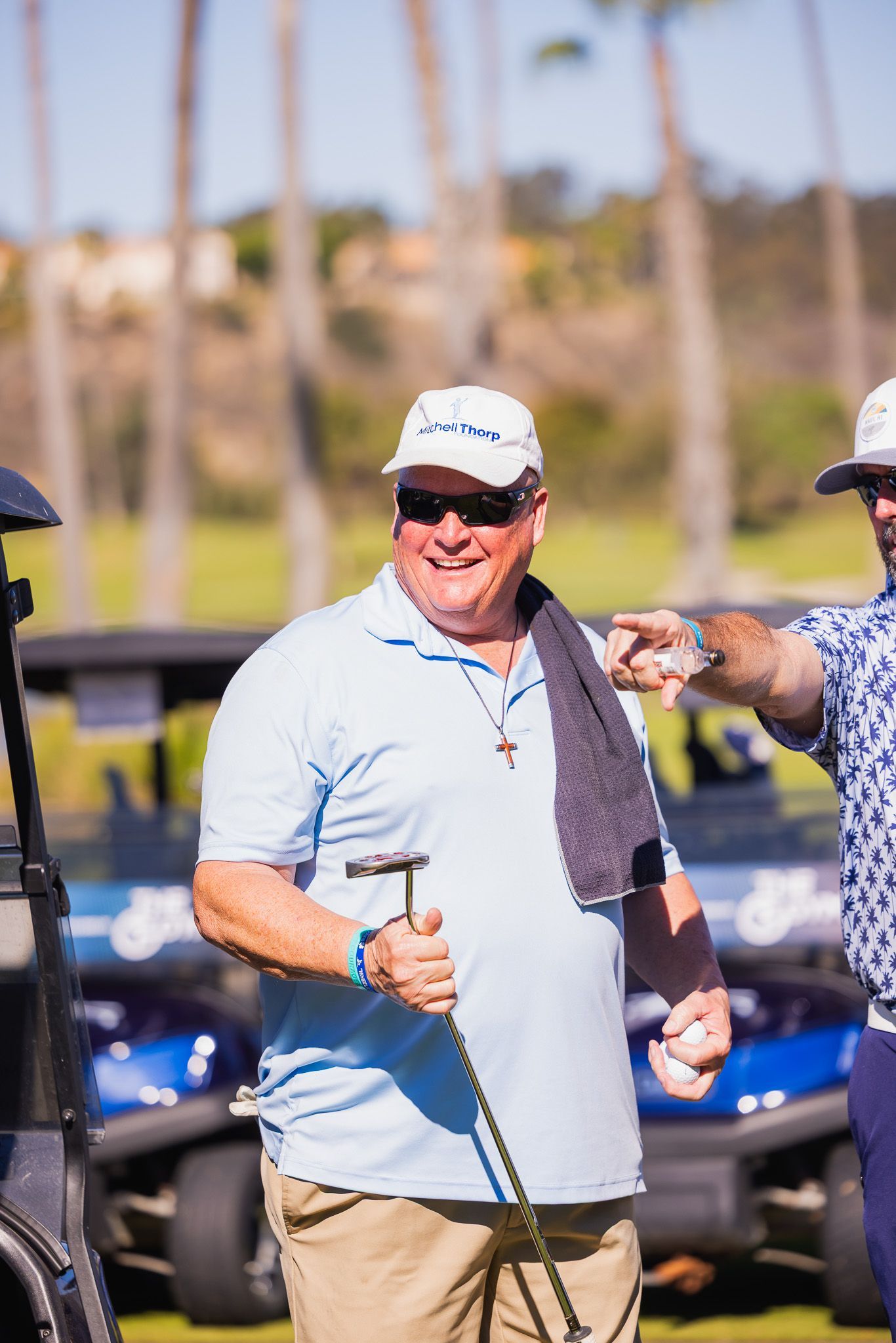 Man in light blue shirt holding golf club, smiling at golf course, with another man pointing.