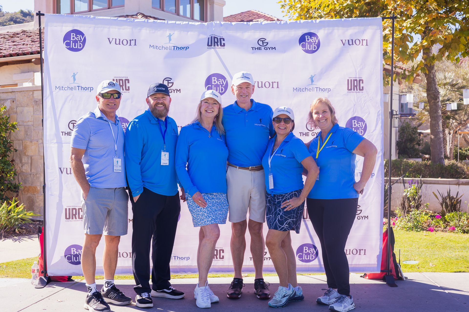 Group of six people in blue shirts and hats posing in front of a banner at an outdoor event.