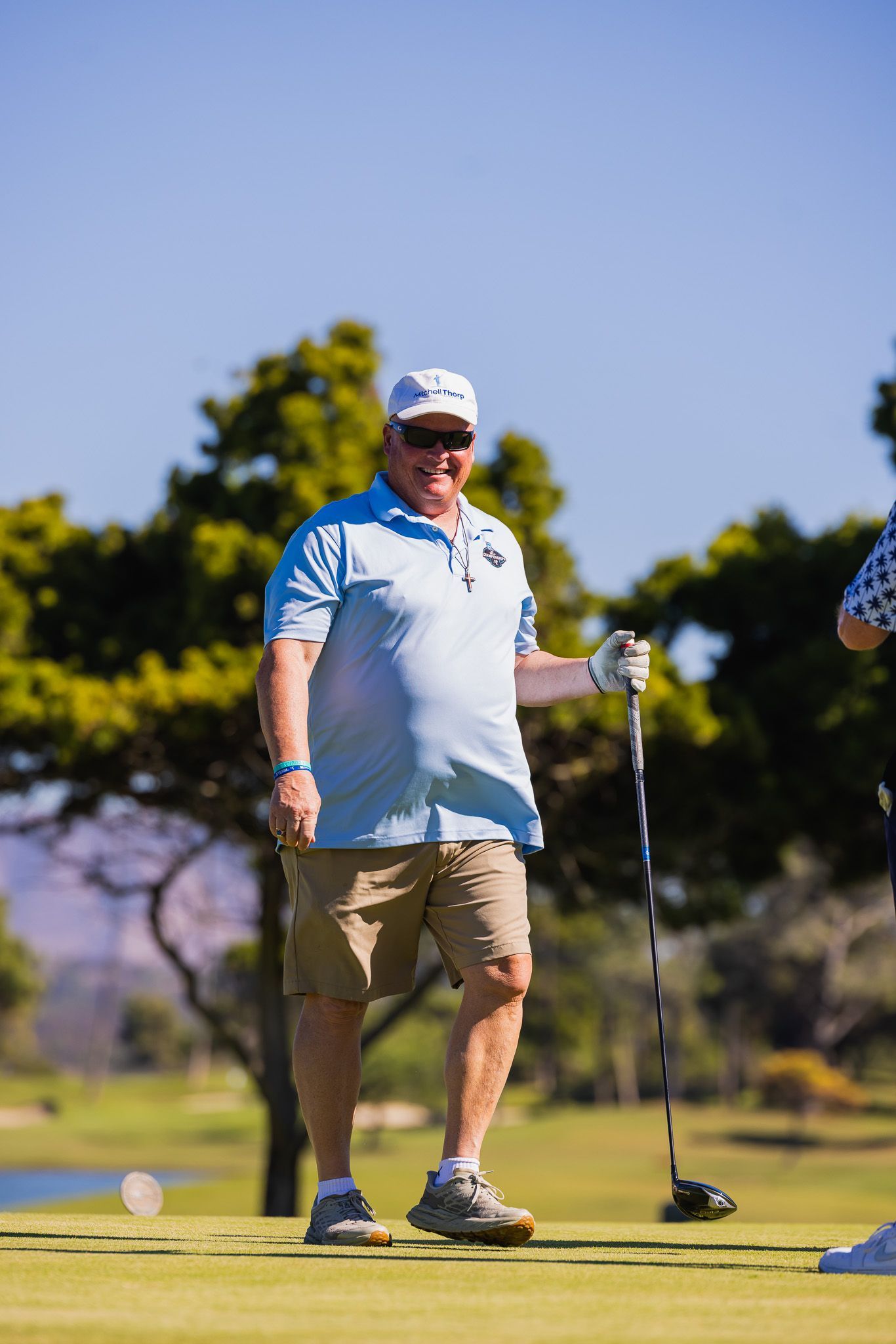 Man in light blue polo and khaki shorts holding a golf club on a green, sunny day.