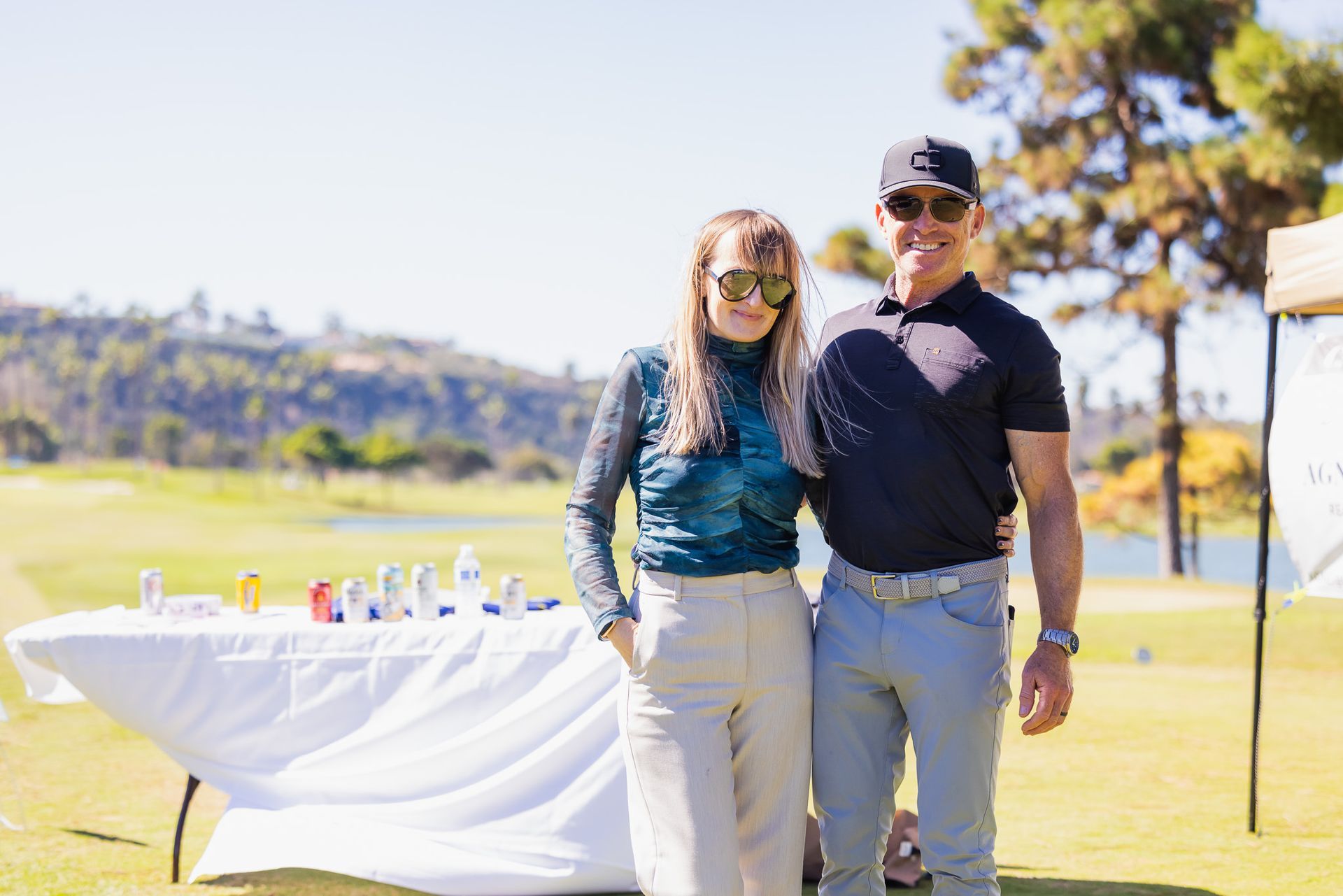 Woman and man pose on a golf course, sunny day. He wears a cap and sunglasses. Table of drinks visible.