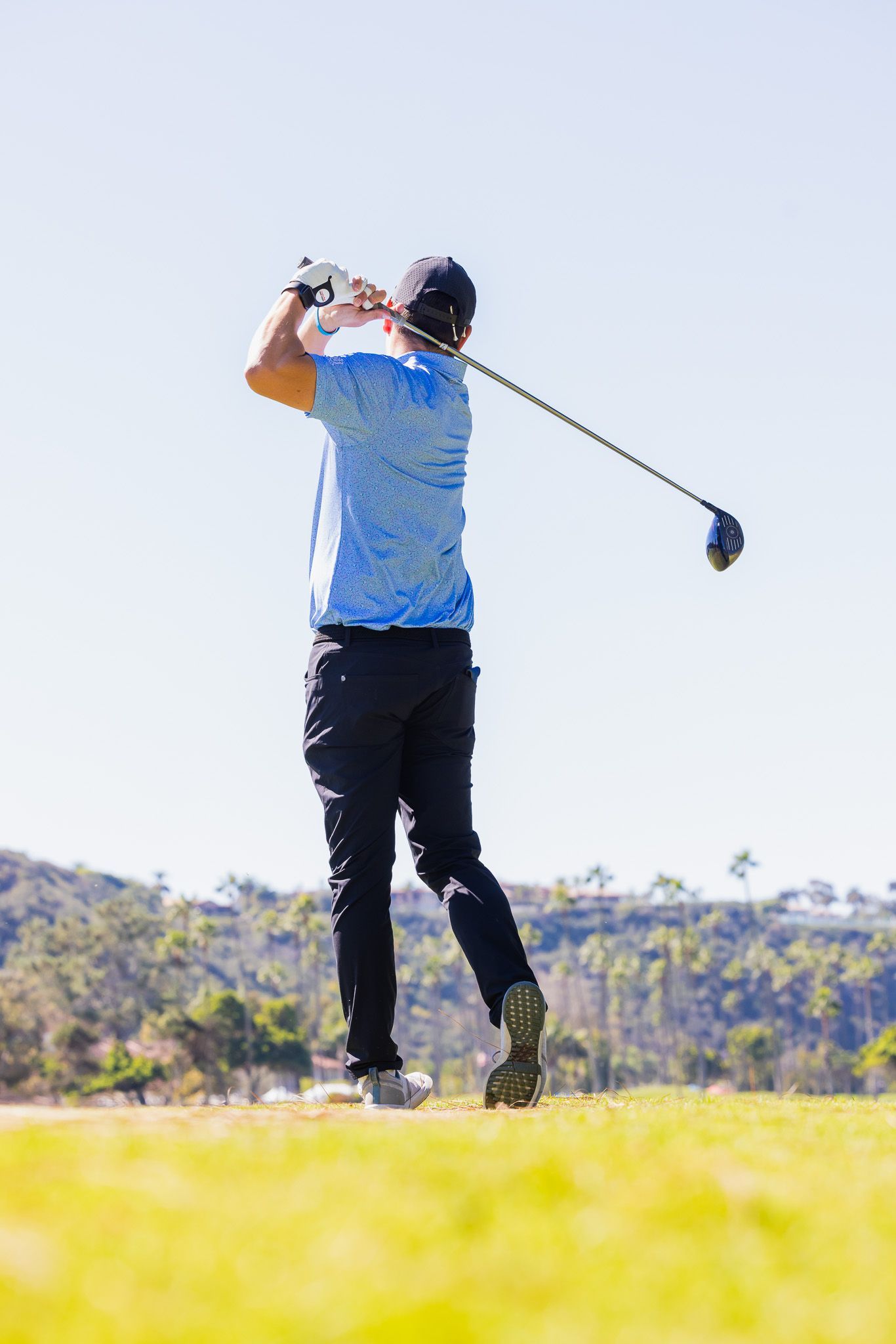 Golfer swings a club on a sunny golf course, wearing a blue shirt and black pants.