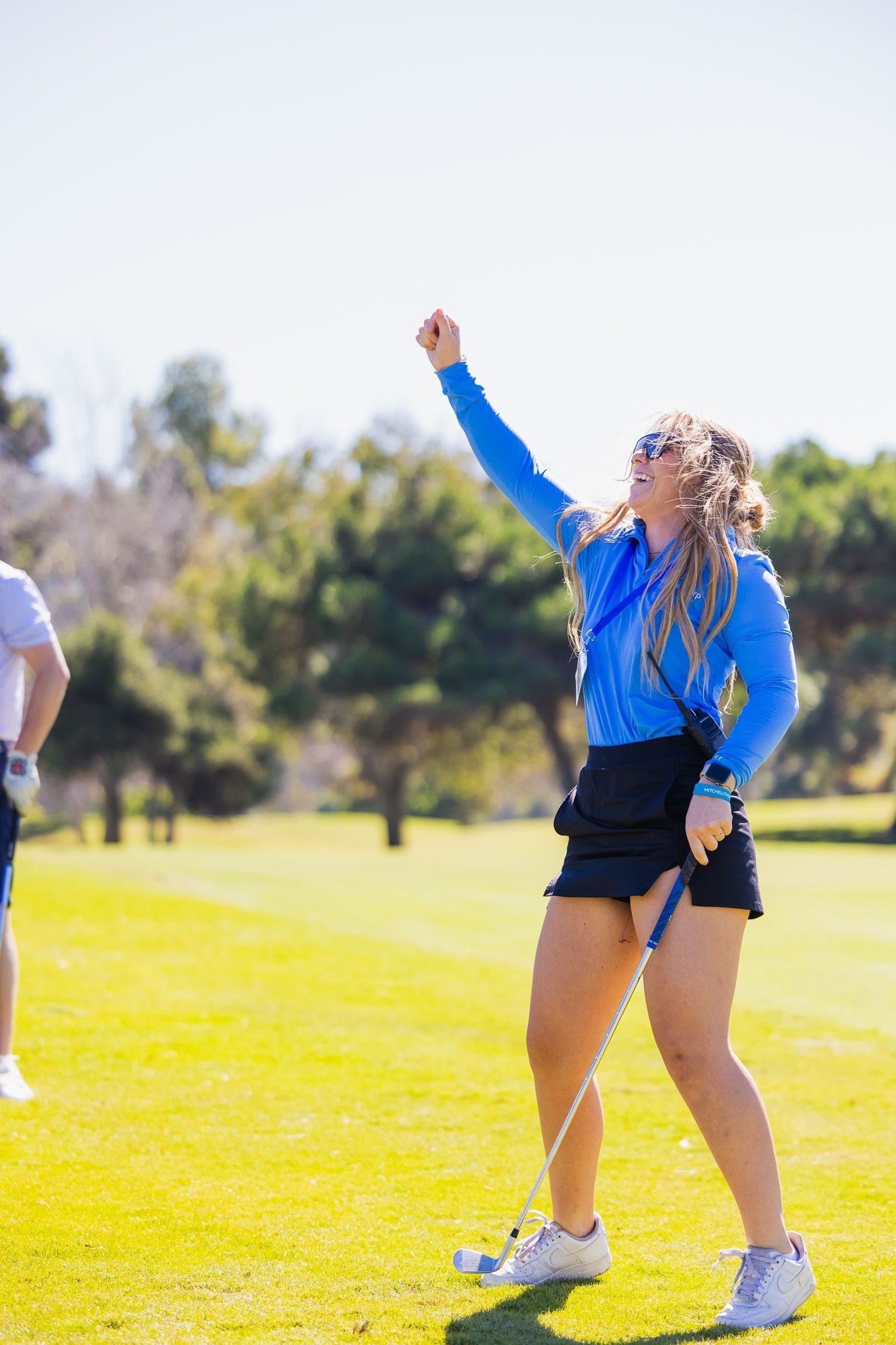 Woman in blue cheering on a golf course, raising arm. Wearing athletic clothing, sunny day.