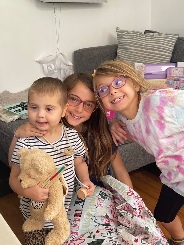 Three children smiling, one holding a teddy bear. They are indoors near a couch and blankets.