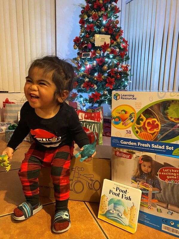Excited child with gifts by Christmas tree, laughing joyfully, wearing plaid pants and holding toys.