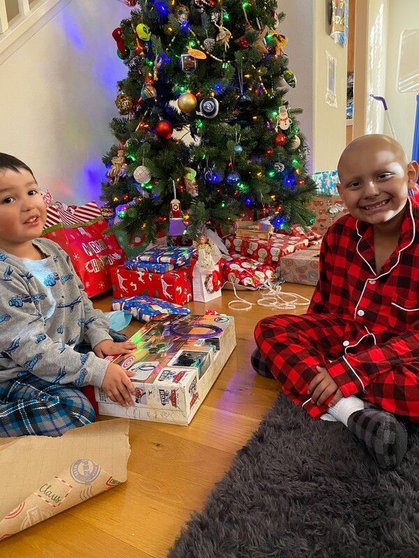 Two children smile, sitting near a decorated Christmas tree and wrapped presents. One child wears pajamas.