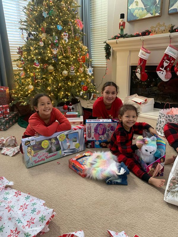 Three smiling children by Christmas tree, surrounded by wrapped gifts. One child is holding a stuffed animal.