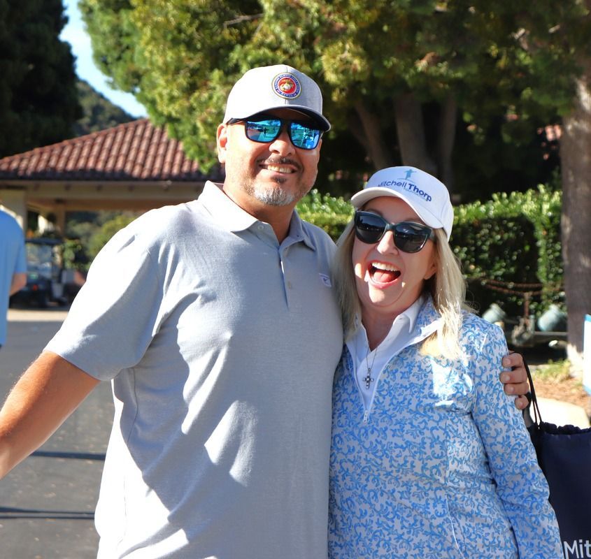 Man and woman smiling, embracing outdoors. Man wears gray polo, hat, sunglasses. Woman in blue floral shirt, hat, sunglasses.