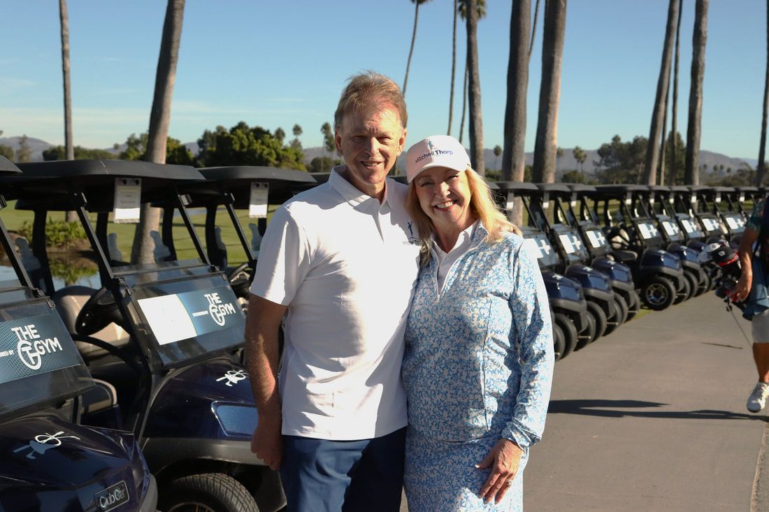 Couple smiling, posing near golf carts on a sunny day. Palm trees and golf course visible.