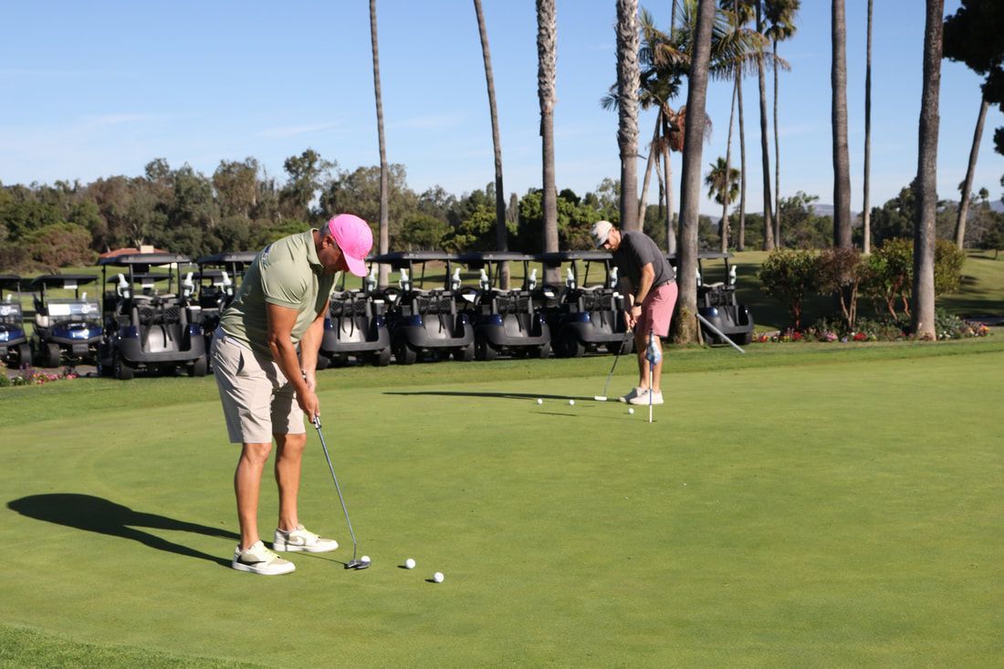 Two men putting golf balls on a green. One wears a pink hat, the other has pink shorts. Golf carts in the background.