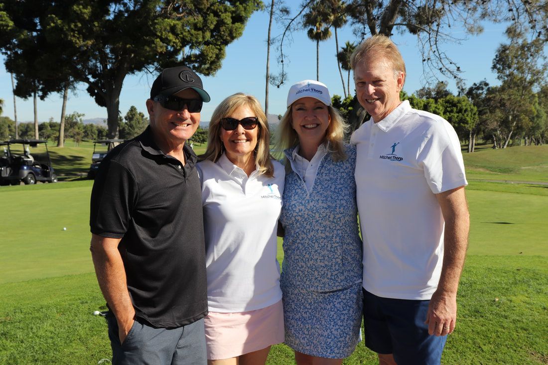 Four people smiling on a golf course. Two women in golf attire, two men in casual wear, sunny day.