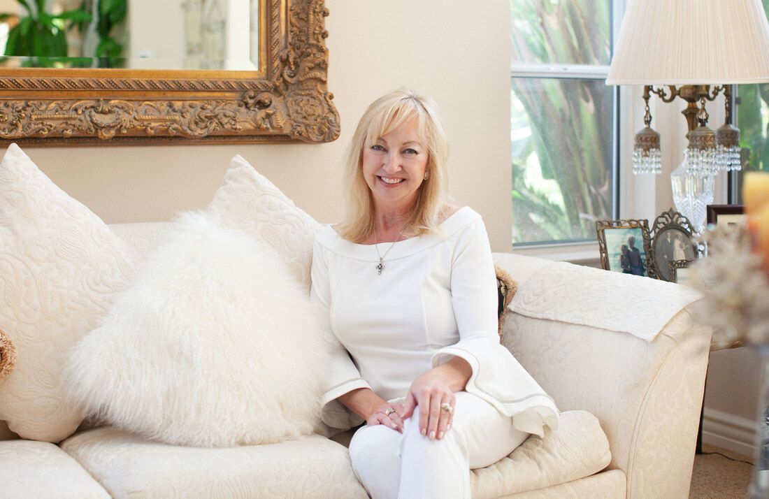 Blonde woman in white outfit smiles on a white sofa in a bright room.