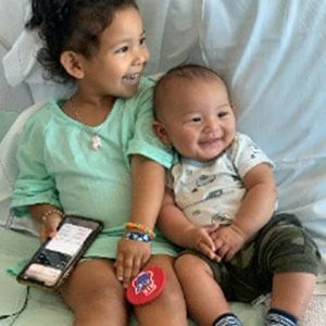 Girl and baby smiling, sitting together. Girl holds phone, baby wears camo pants, both on a bed.