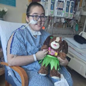 Woman in hospital gown, glasses, with stuffed dog, in hospital room.