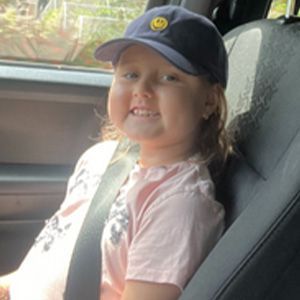 Young girl in a car, smiling, wearing a blue cap with a smiley face.