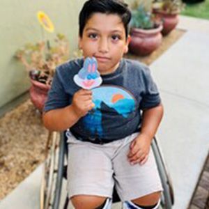 Young boy in a wheelchair holding a Bugs Bunny popsicle; wearing a gray shirt, tan shorts, outside.