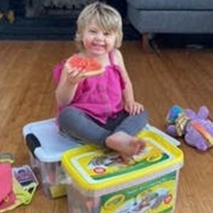 Young girl in pink top, holding watermelon, sits on storage containers.