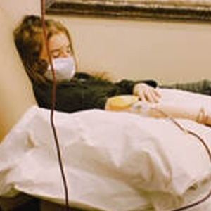 Woman in mask and dark top receiving IV treatment, resting on a bed.