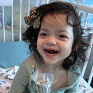 Smiling young girl with dark curly hair and a bow, in a hospital bed.