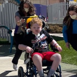 Boy in wheelchair outdoors, pushed by a person; another person walks nearby. All wear masks.