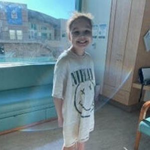 Young person wearing a Nirvana t-shirt, smiling, in a room with a window and a cabinet.